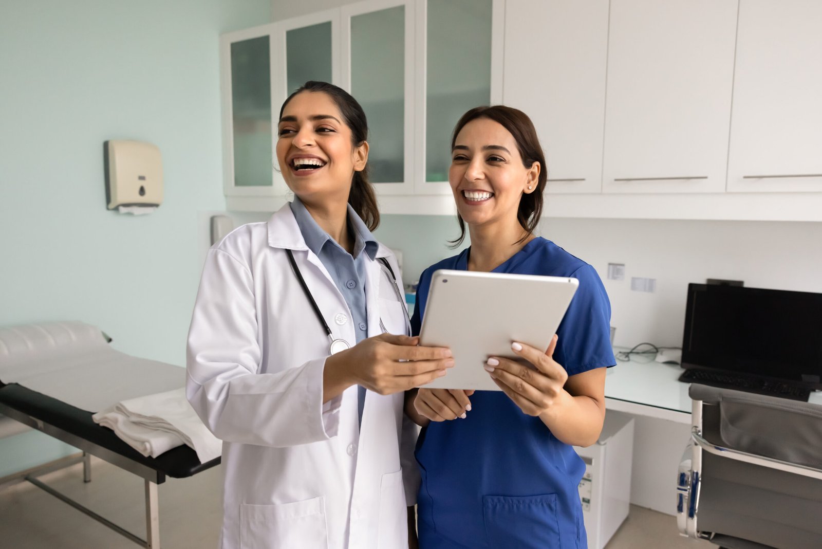 Two happy diverse female doctor colleagues using digital tablet in hospital exam room, looking away off shot, smiling, laughing, standing close. Indian practitioner consulting surgeon